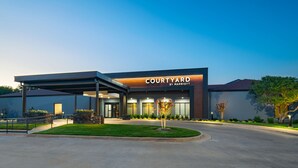 Une entrée d’hôtel moderne avec une allée couverte, une pelouse bien entretenue et une enseigne indiquant « Courtyard by Marriott ».
