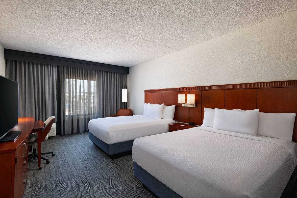 A hotel room with two beds, a desk, a TV, and a large window with curtains.