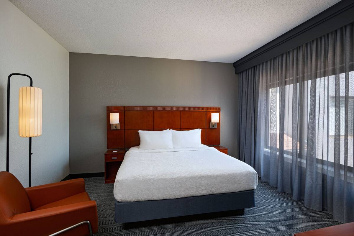 A hotel room with a large bed, a nightstand, a lamp, and a view of the outdoors through sheer curtains.