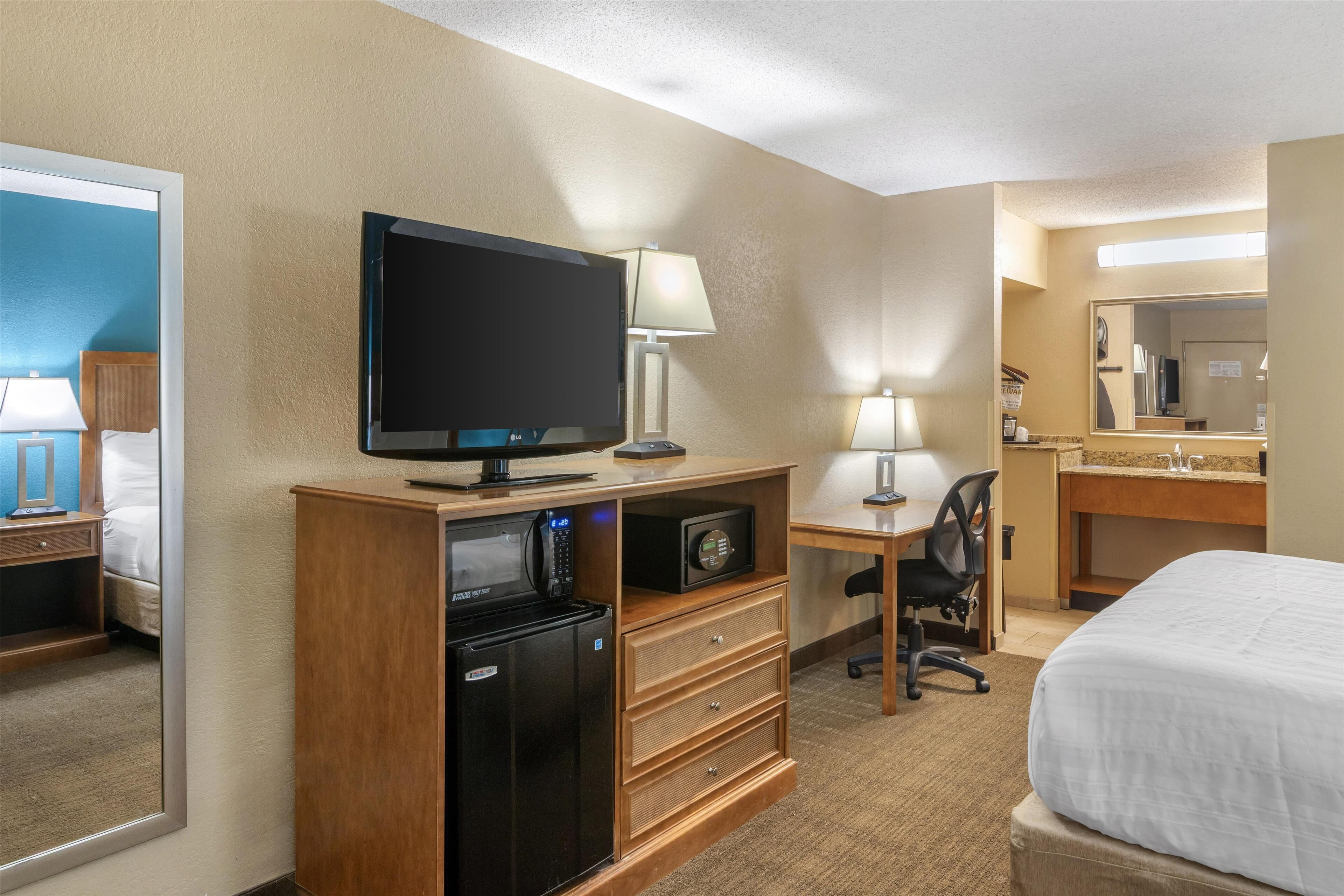 Habitación de hotel con cama, escritorio con computadora, microondas, refrigerador, televisor y espejo.