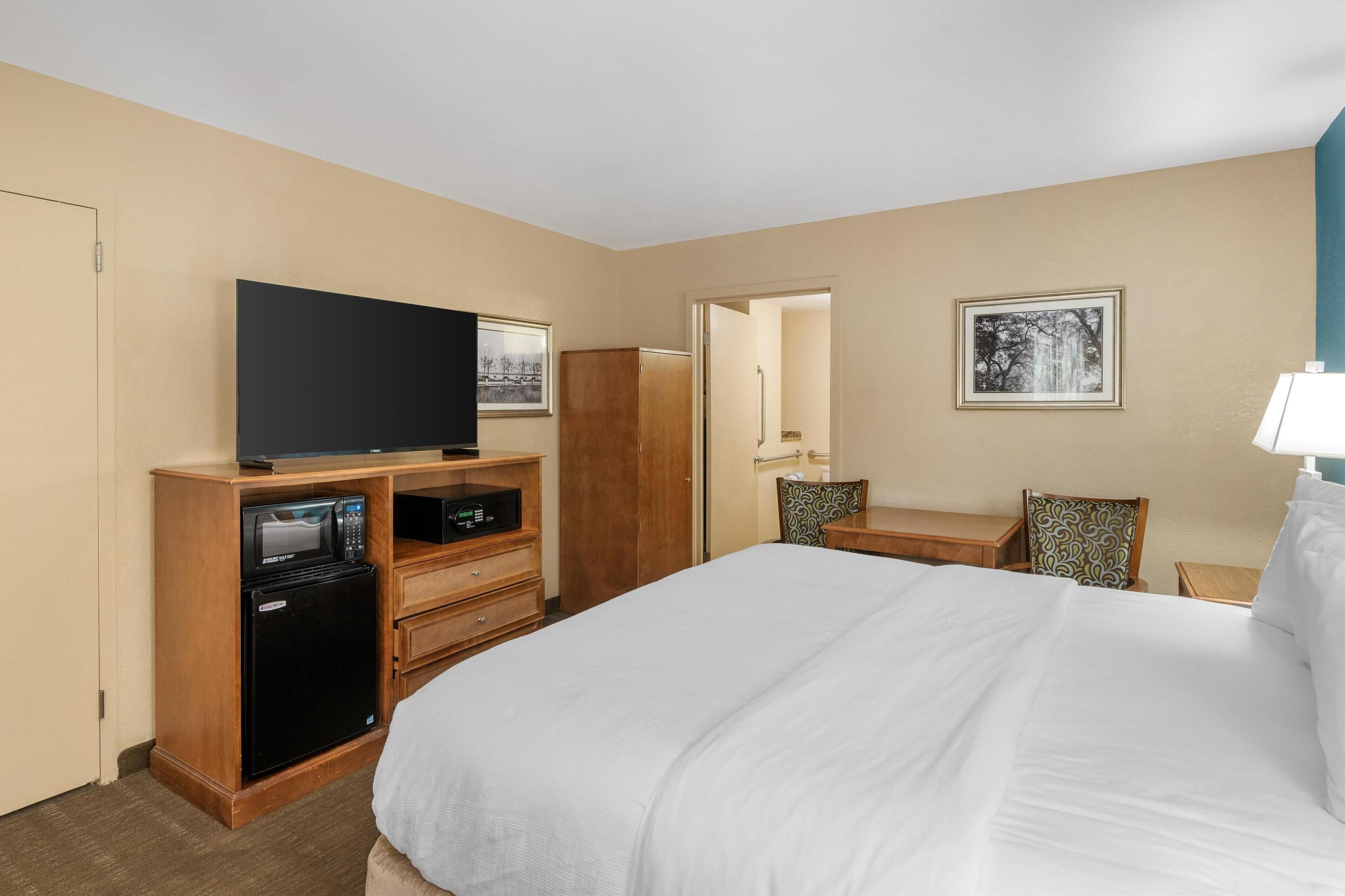 A hotel room with a large bed, a TV on a wooden stand, a microwave, a refrigerator, a desk with a chair, and a framed picture on the wall.
