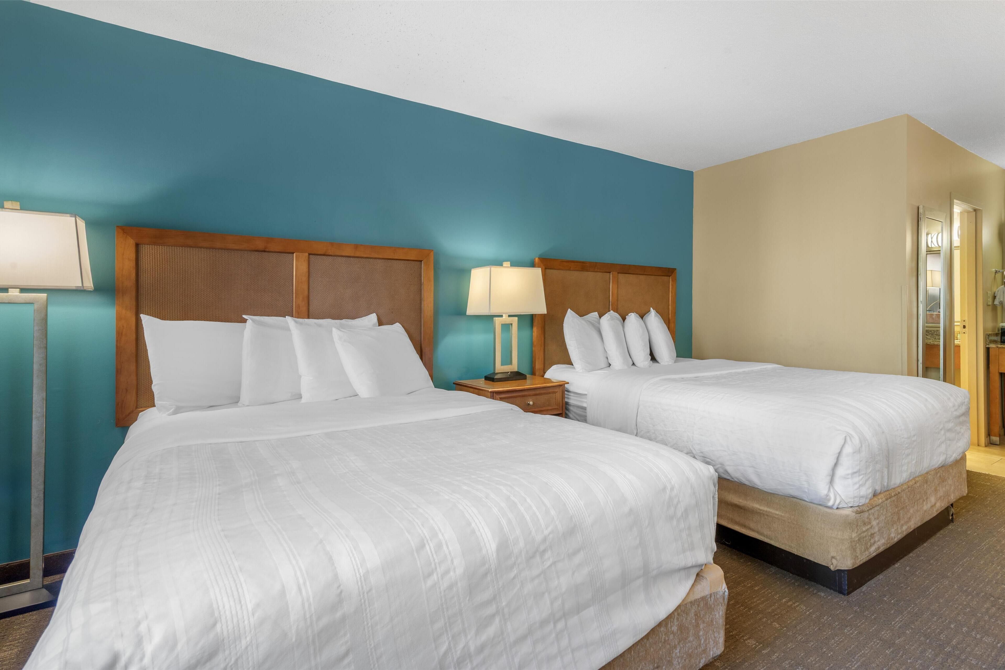 Quarto de hotel com duas camas, cada uma com roupa de cama branca, cabeceira de madeira e mesa de cabeceira com abajur.