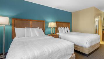 Quarto de hotel com duas camas, cada uma com roupa de cama branca, cabeceira de madeira e mesa de cabeceira com abajur.