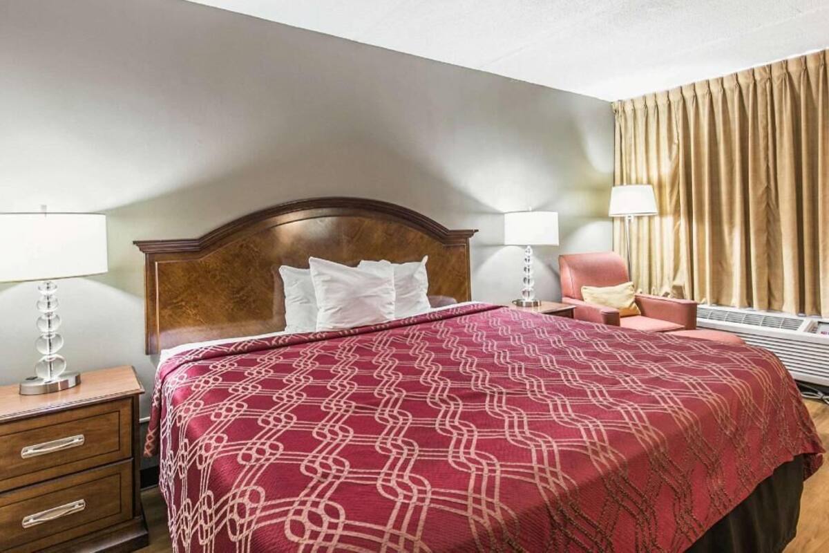 A hotel room with a large bed, two bedside tables with lamps, a chair, a window with curtains, and a wall-mounted heater.