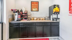 Una barra de desayuno con máquinas de café, un dispensador de zumo de frutas y diferentes opciones para el desayuno.