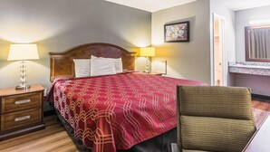 Una habitación de hotel con cama, mesita de noche, lámpara, silla y un cuadro en la pared.