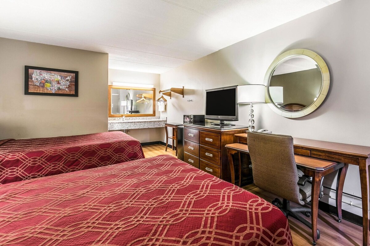 Habitación de hotel con dos camas, un escritorio, una silla, un televisor, un espejo y un baño visible al fondo.