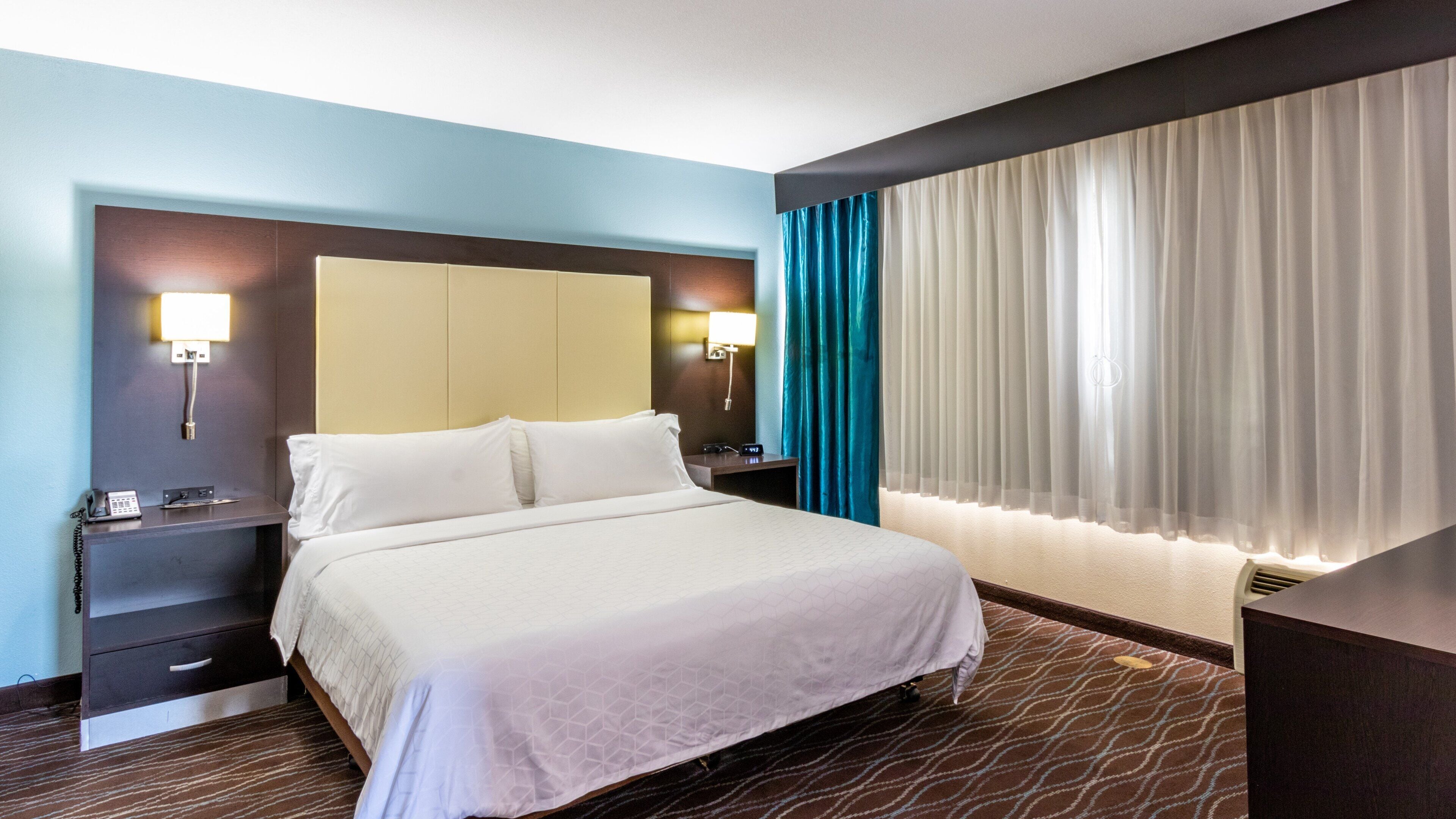 A hotel room with a bed, bedside tables, a desk, a phone, and a window with curtains.