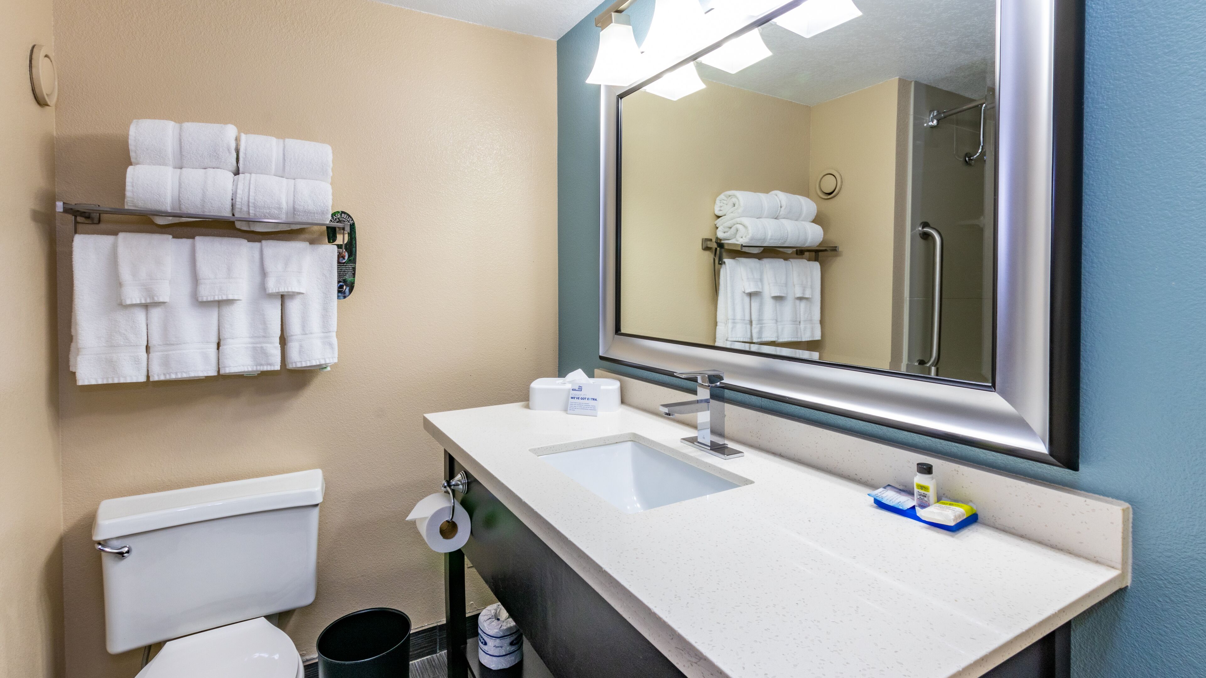 A bathroom with a toilet, sink, and mirror. There are towels hanging on a rack and a trash can.