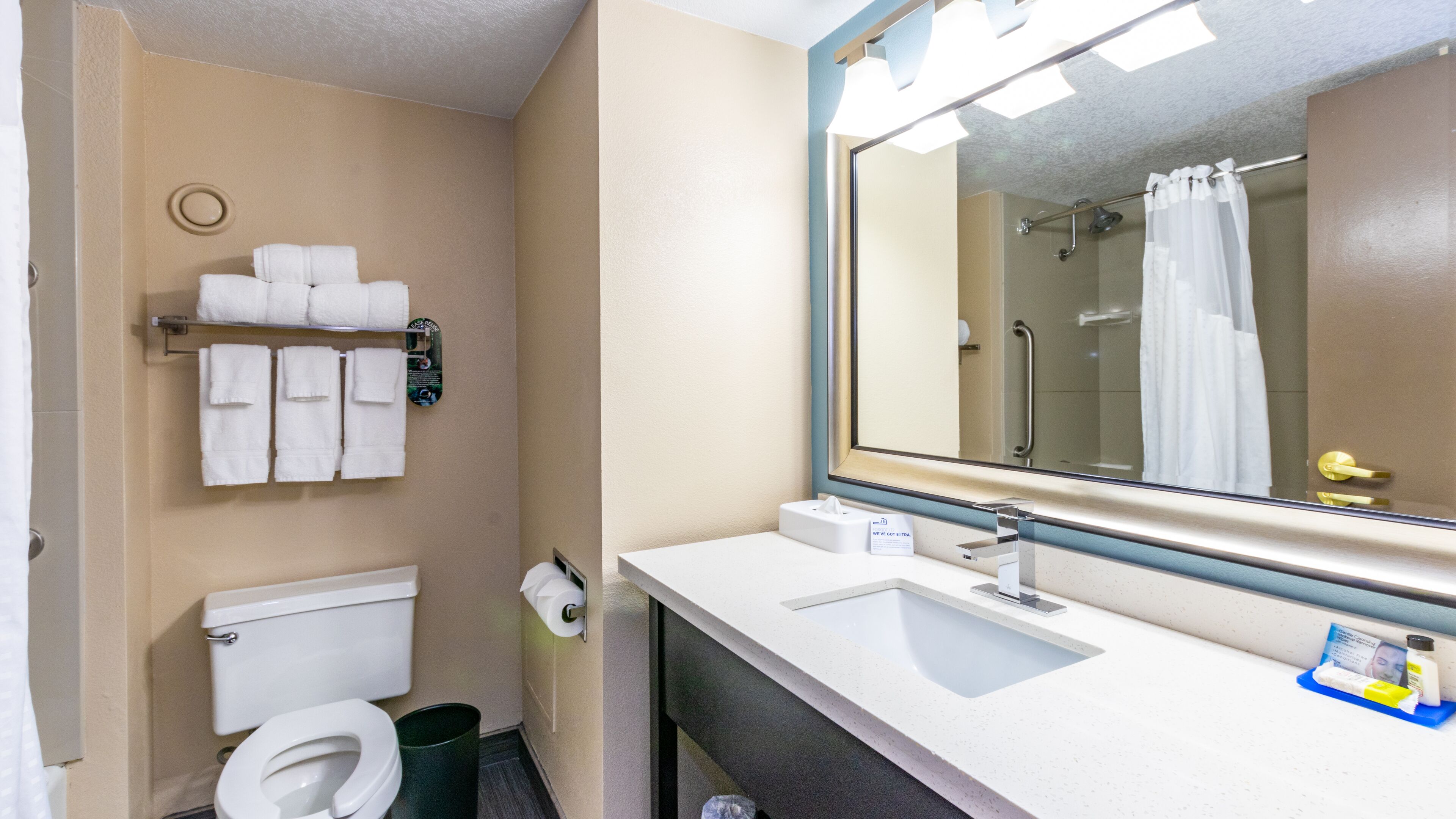 A bathroom with a toilet, sink, and mirror. There are towels hanging on the wall and a trash can.