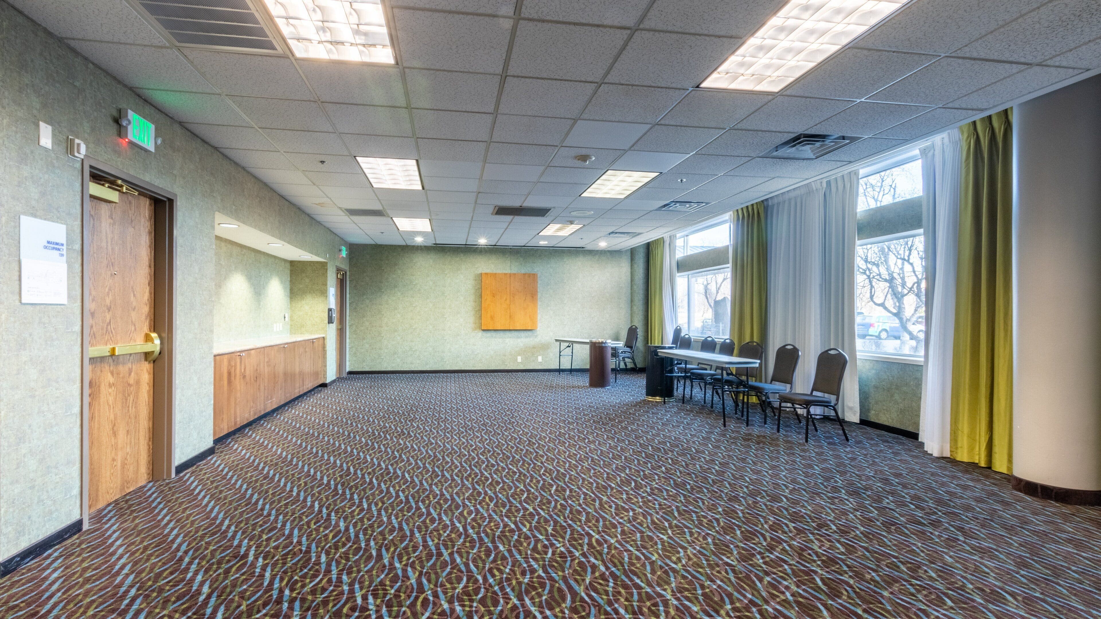 Un salón de conferencias con alfombra estampada, una mesa grande con sillas, un cuadro en la pared y ventanas con cortinas.