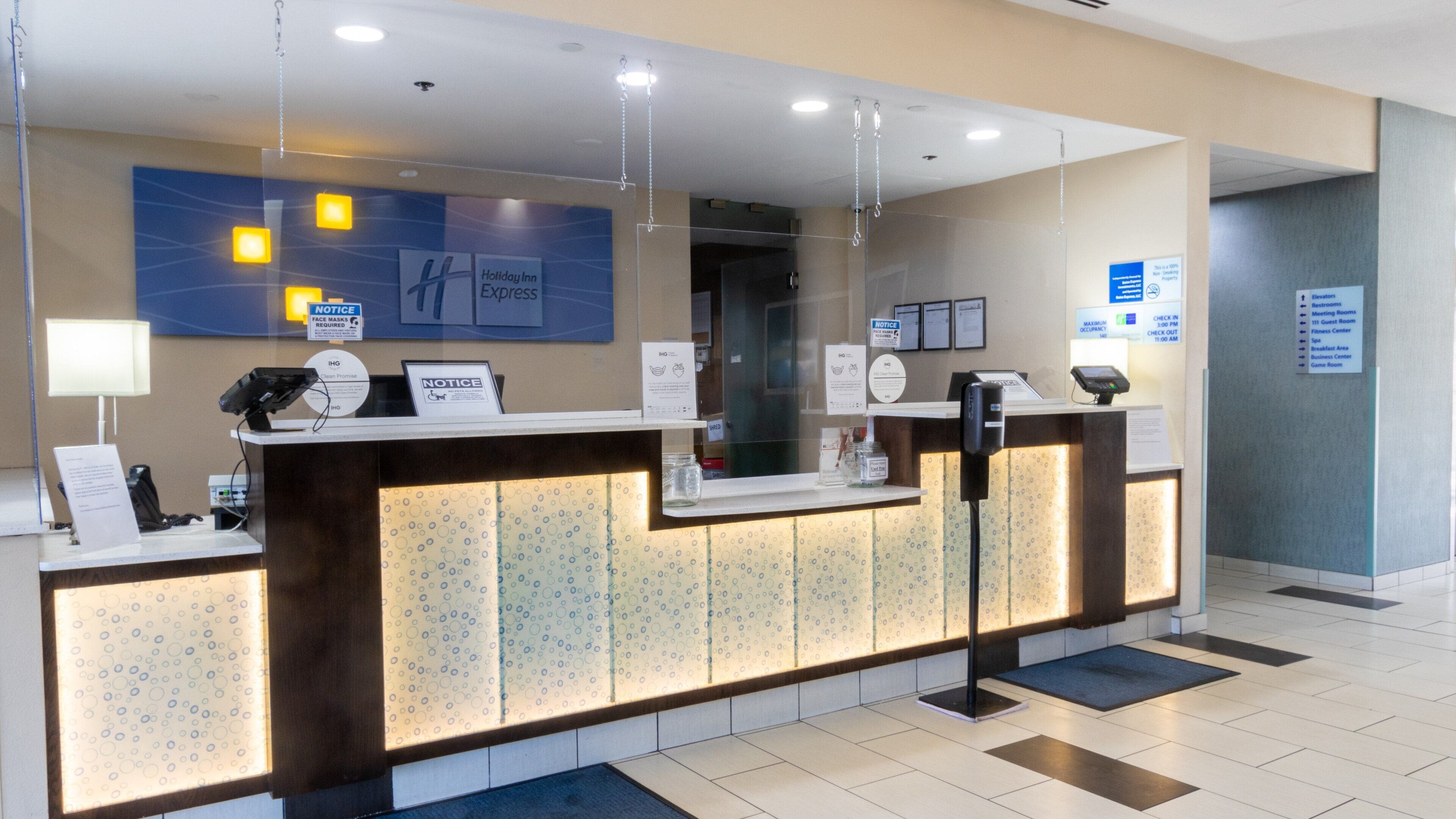 Una recepción de hotel con una estación de registro digital, un separador de vidrio y carteles informativos.