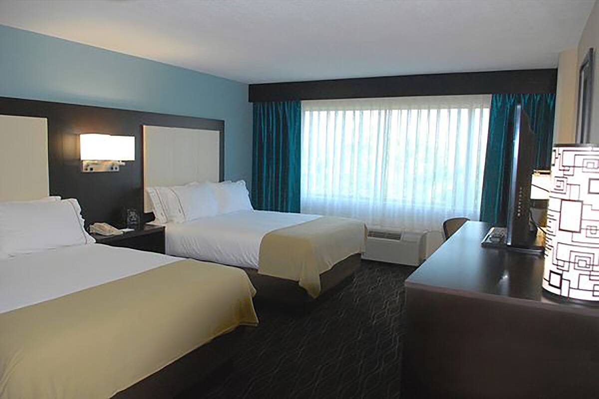 Habitación de hotel con dos camas, un escritorio, una silla, una lámpara y un ventanal grande con cortinas.