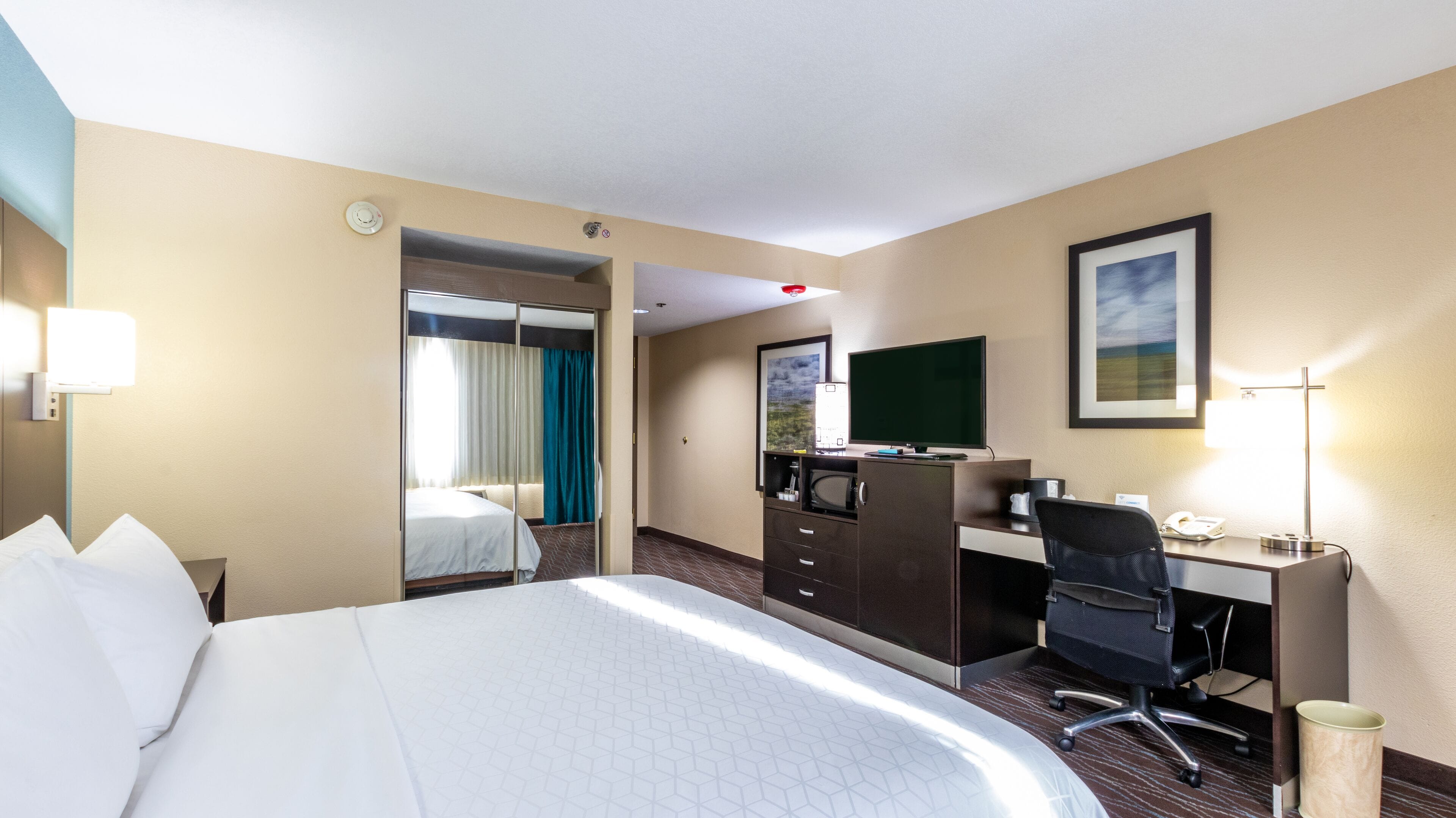 A hotel room with a large bed, a desk with a computer, a chair, a TV, and a mirror.