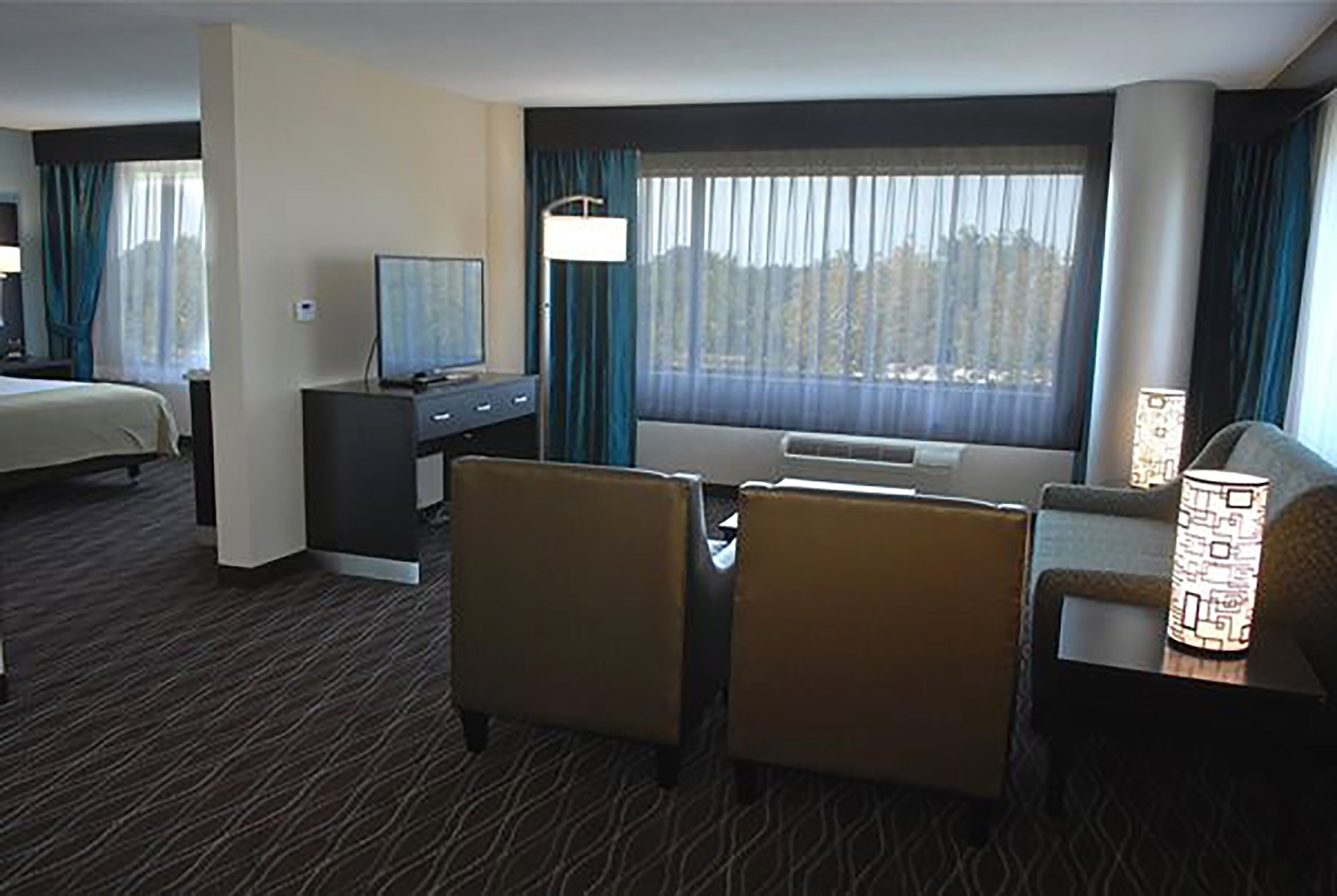 A hotel room with a bed, a desk with a television, two armchairs, a sofa, a window with curtains, and a lamp.