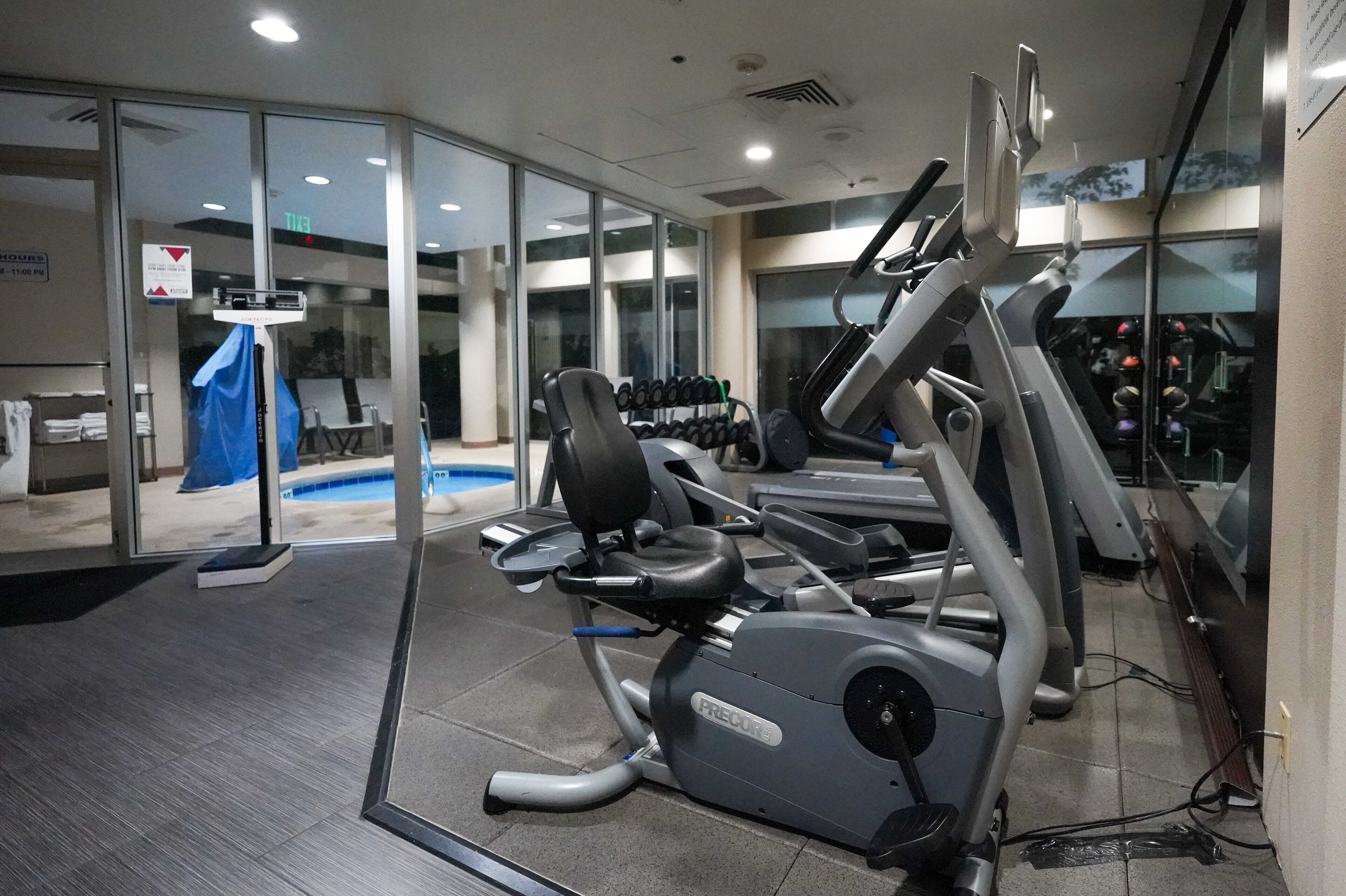 A gym with cardio machines, weight benches, and a glass wall overlooking a pool area.