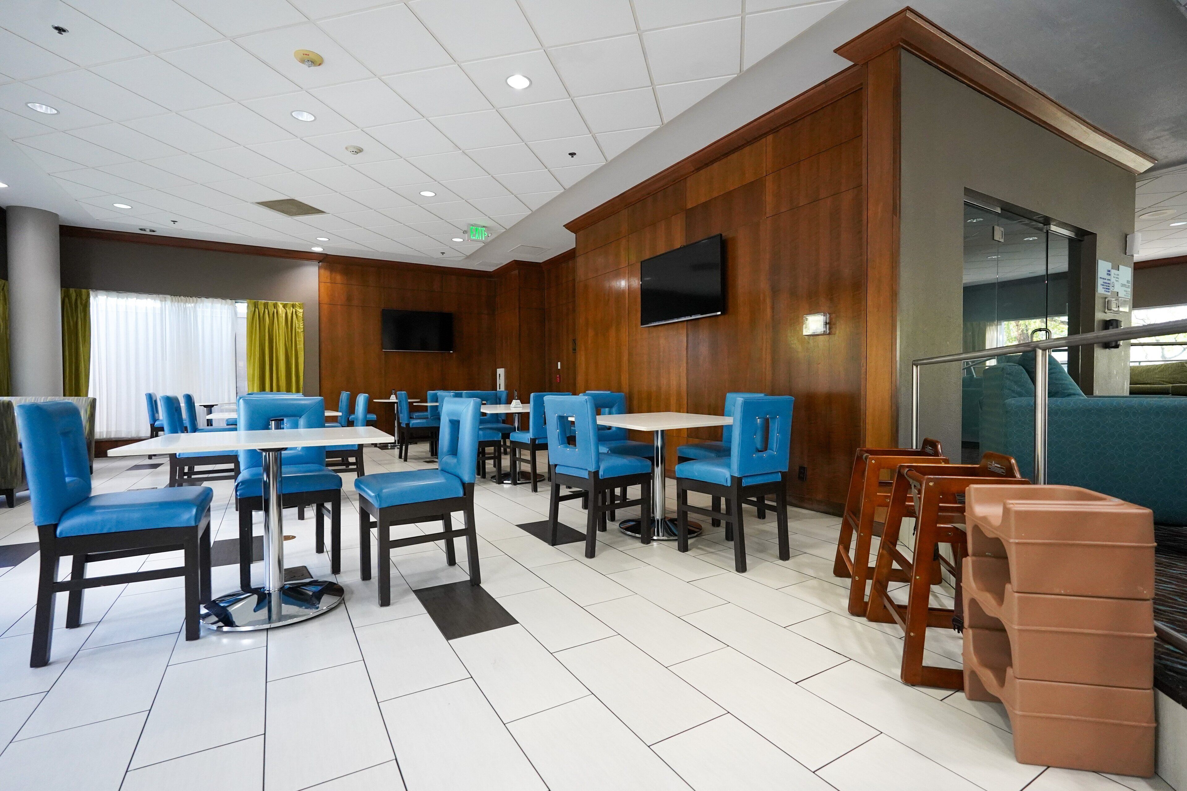 A hotel dining area with blue chairs, white tables, and a wooden wall with a TV.