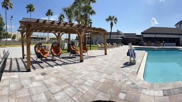 Una piscina con sillones de descanso, una pérgola y un edificio al fondo.