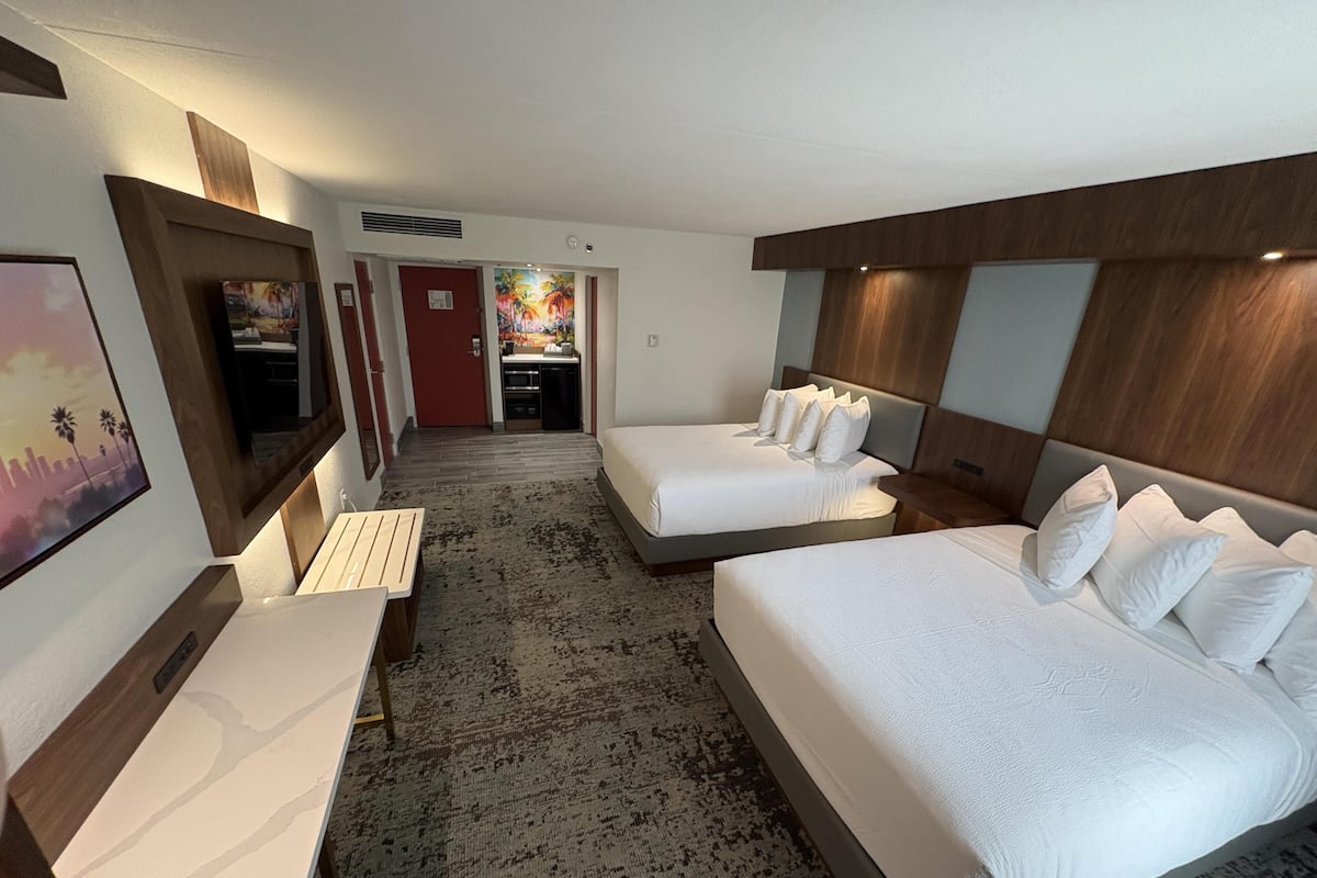 A hotel room with two beds, a desk, a TV, and a view of the kitchen and dining area.