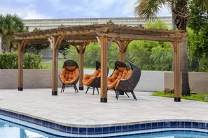 Un espace aménagé au bord de la piscine, avec une pergola, deux fauteuils en osier dotés de coussins et un palmier.