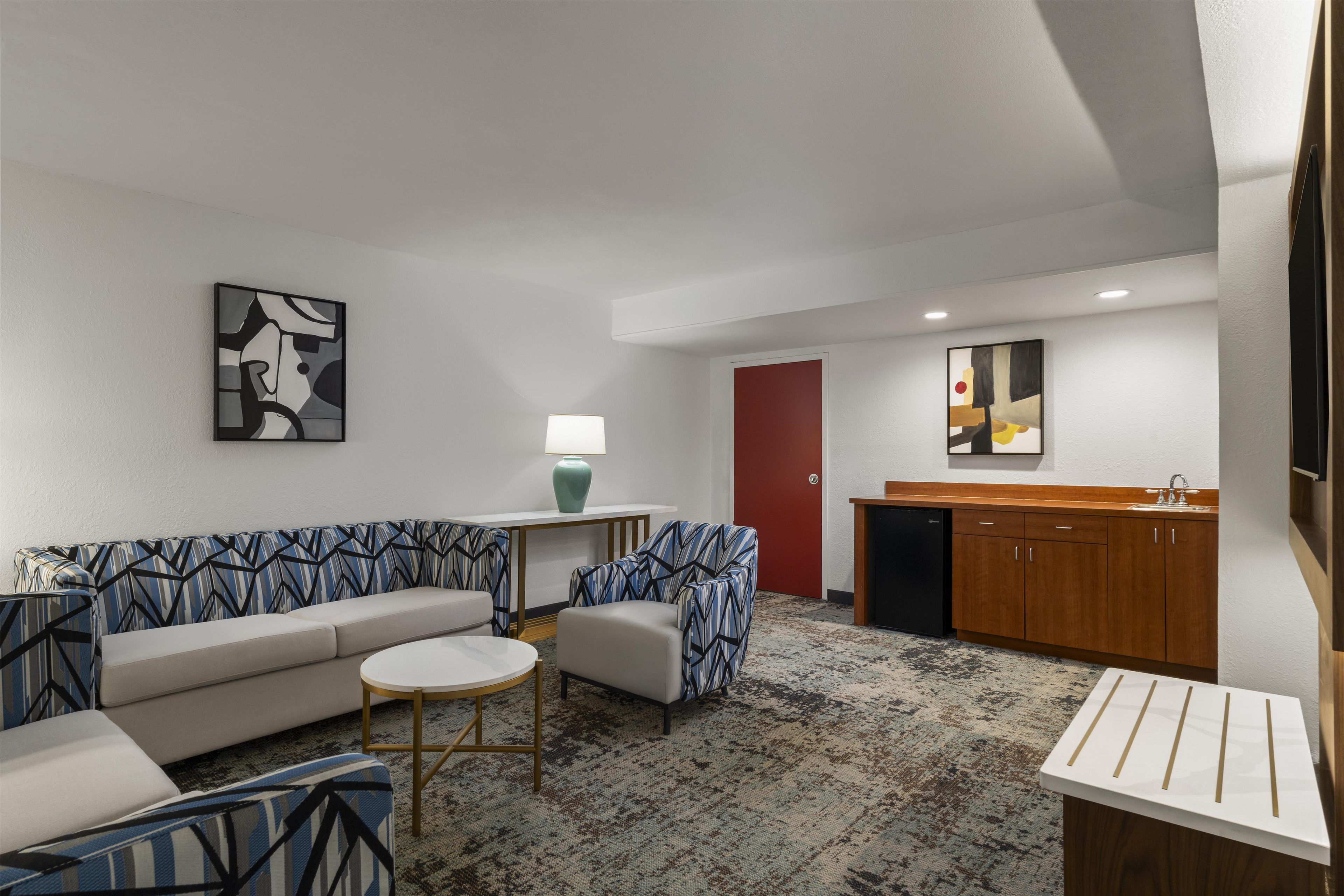 A modern hotel room with a sofa, armchairs, a small table, a lamp, a red door, a kitchen area with a sink, and a cabinet.