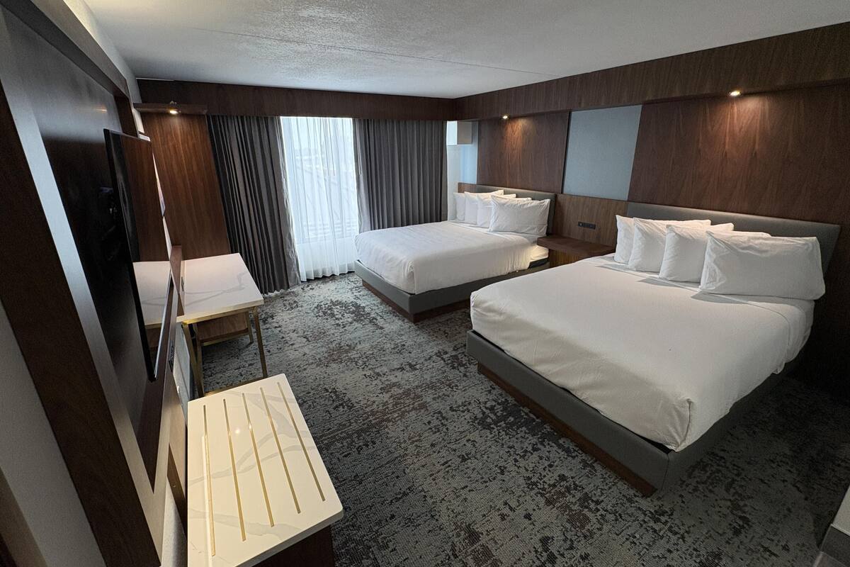 A hotel room with two beds, a desk, a TV, and a large window with curtains.