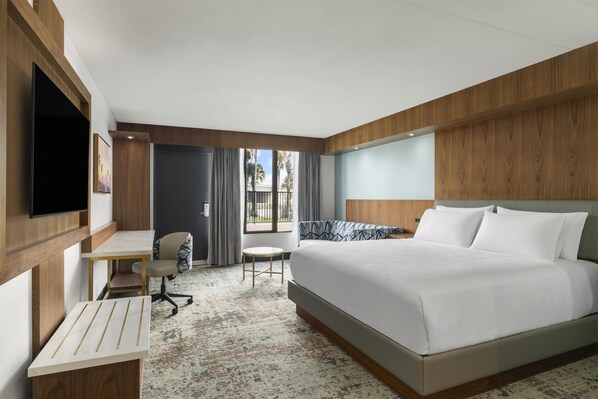A hotel room with a large bed, a desk, a chair, a television, and a view of palm trees through the window.