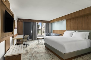 Quarto de hotel com uma cama grande, uma escrivaninha, uma cadeira, televisão e vista para palmeiras pela janela.