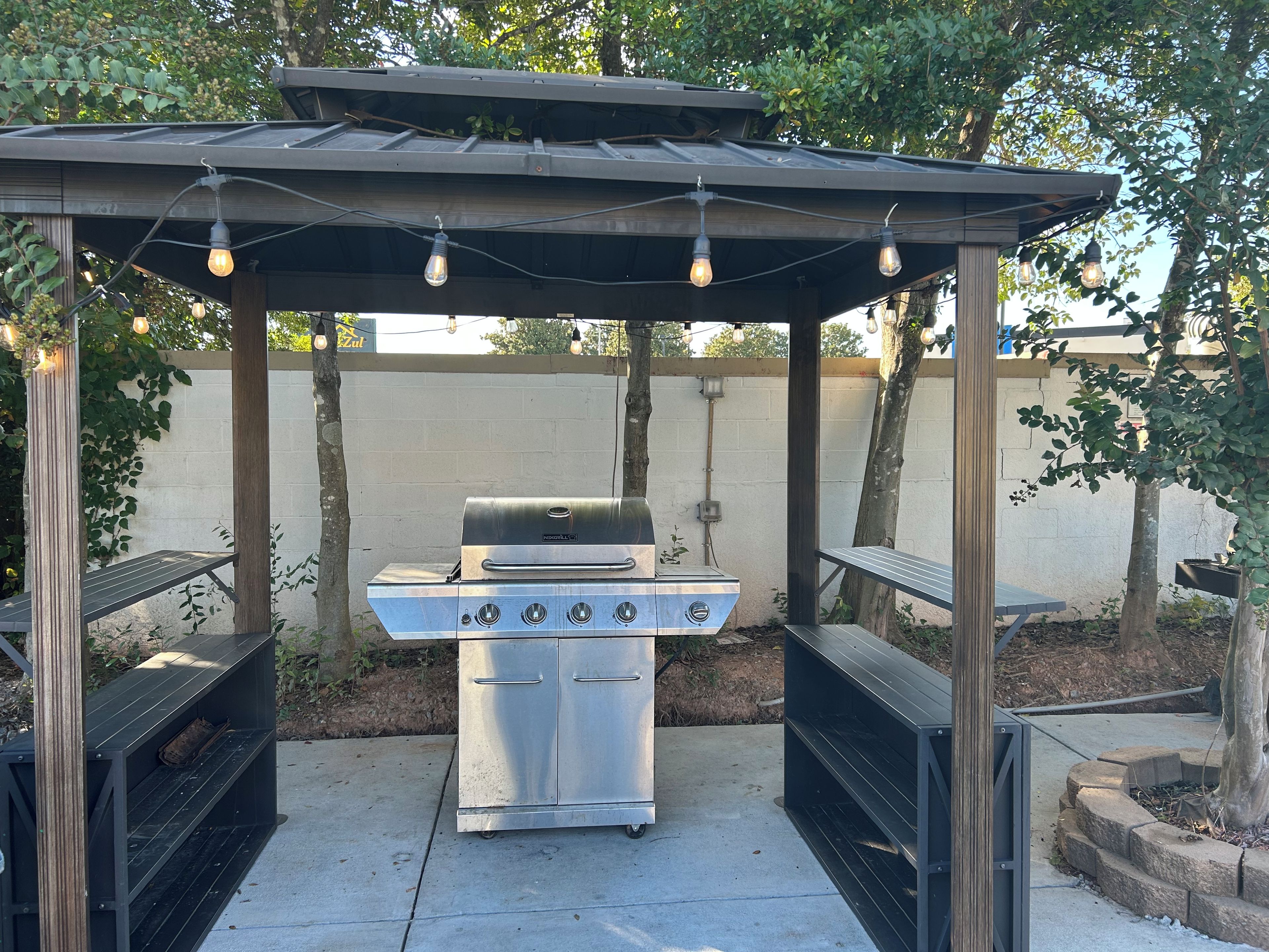 An outdoor barbecue area with a grill, wooden benches, and string lights.