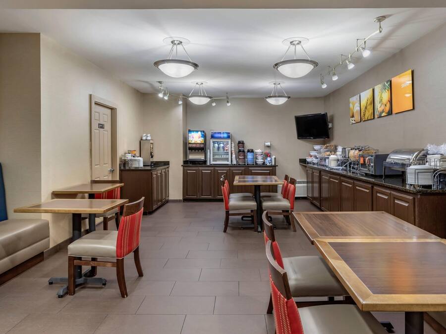 A hotel dining area with multiple tables and benches, a kitchenette, and a television on the wall.