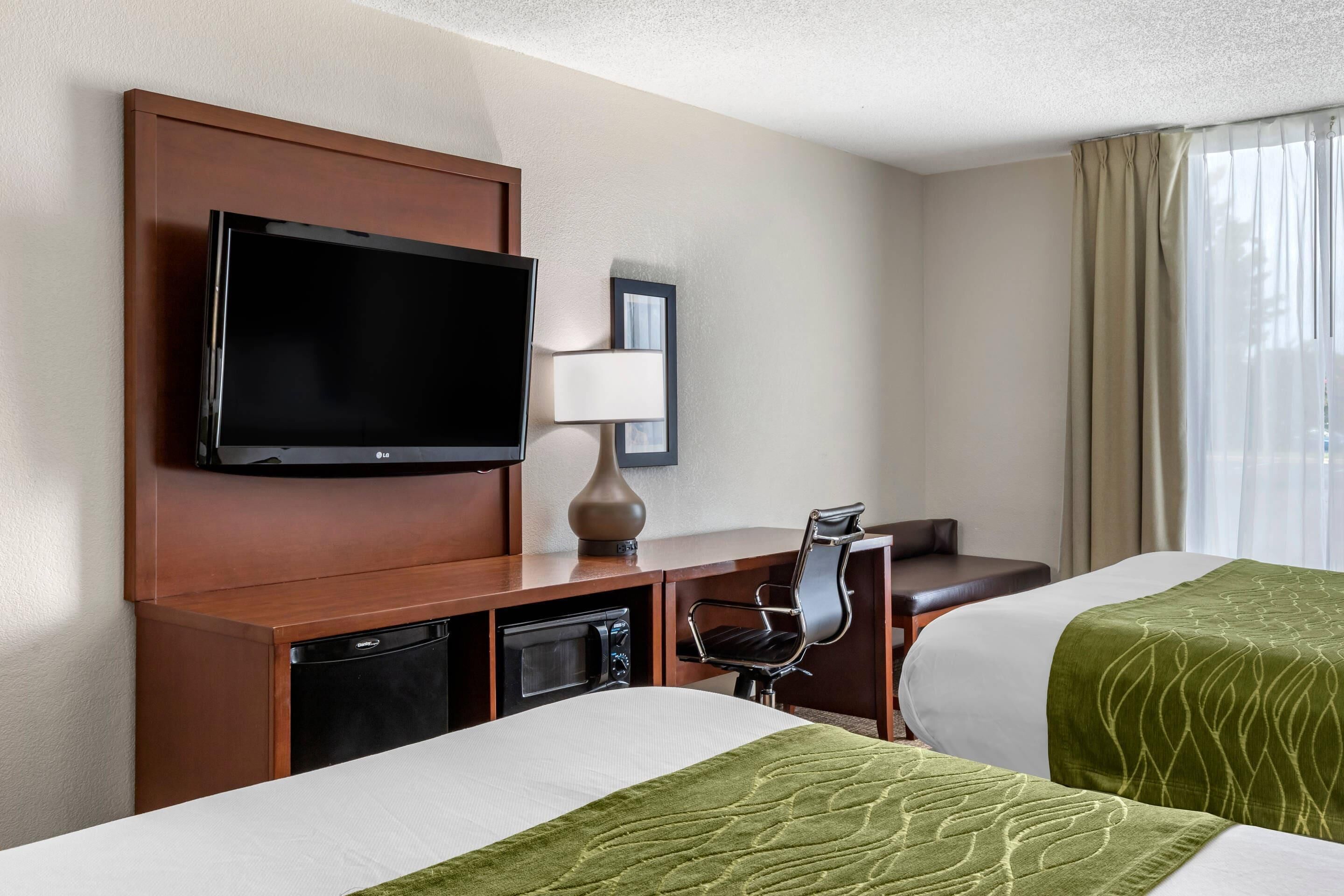 A hotel room with a flat-screen TV, a desk with a lamp, a chair, two beds with green bedspreads, and a window with curtains.