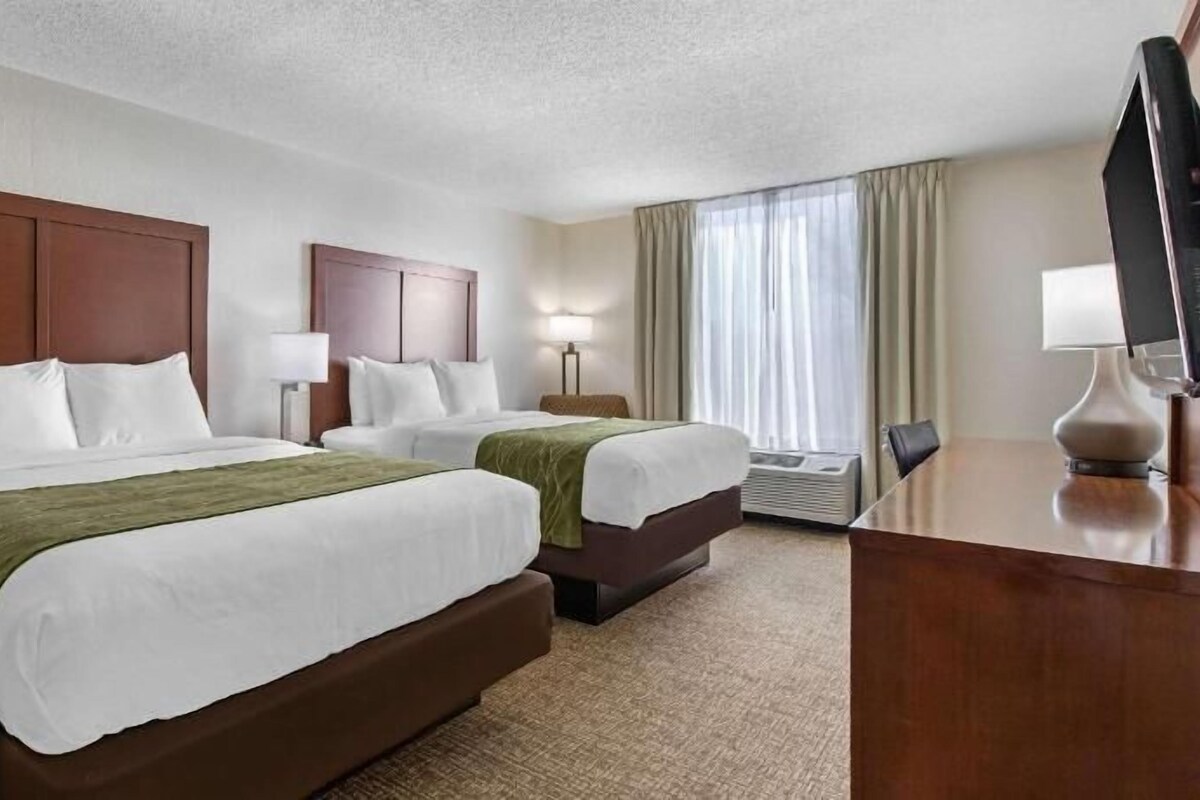 A hotel room with two beds, a desk, a TV, and a window with curtains.