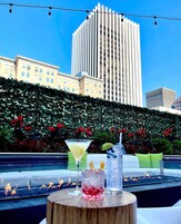 Un bar en la azotea con vista a un edificio alto, un seto decorativo y una piscina.