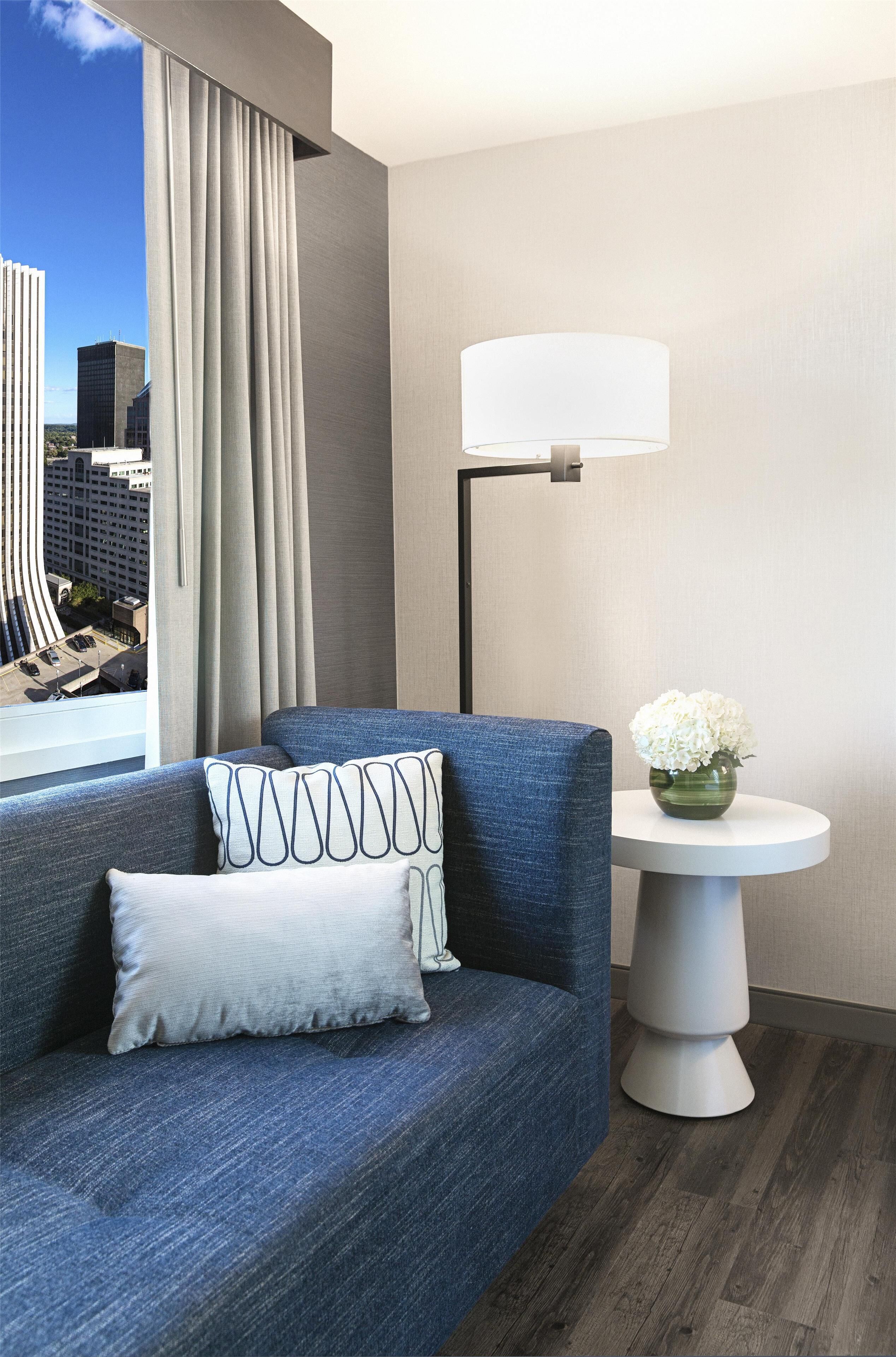 A modern room with a blue sofa, a round table with a vase of flowers, a floor lamp, and a view of a cityscape through large windows.