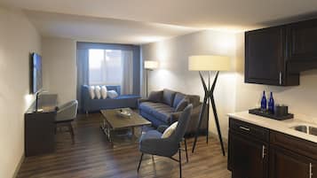 A modern hotel room with a sofa, armchair, dining table, and a kitchen area with cabinets and a sink.