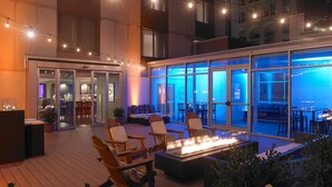 An outdoor seating area with a fire feature, wooden furniture, and a view of the interior through large glass doors. - Hyatt Regency Rochester (Rochester)