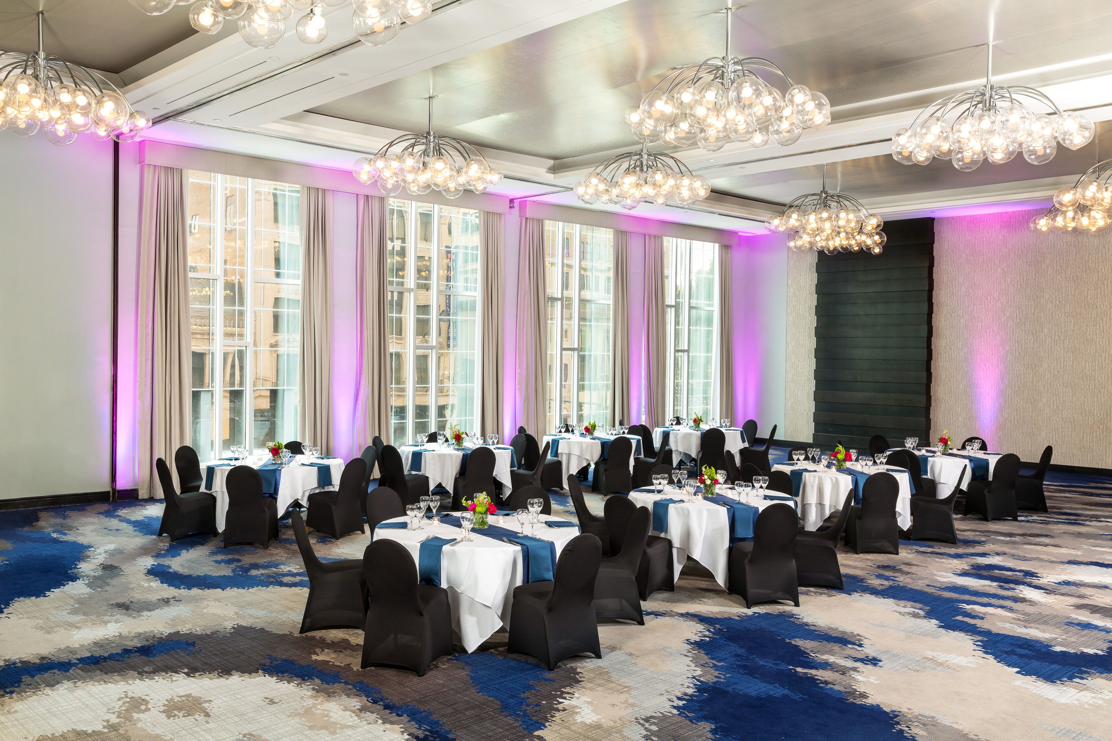 A banquet hall with round tables, black chairs, and chandeliers.