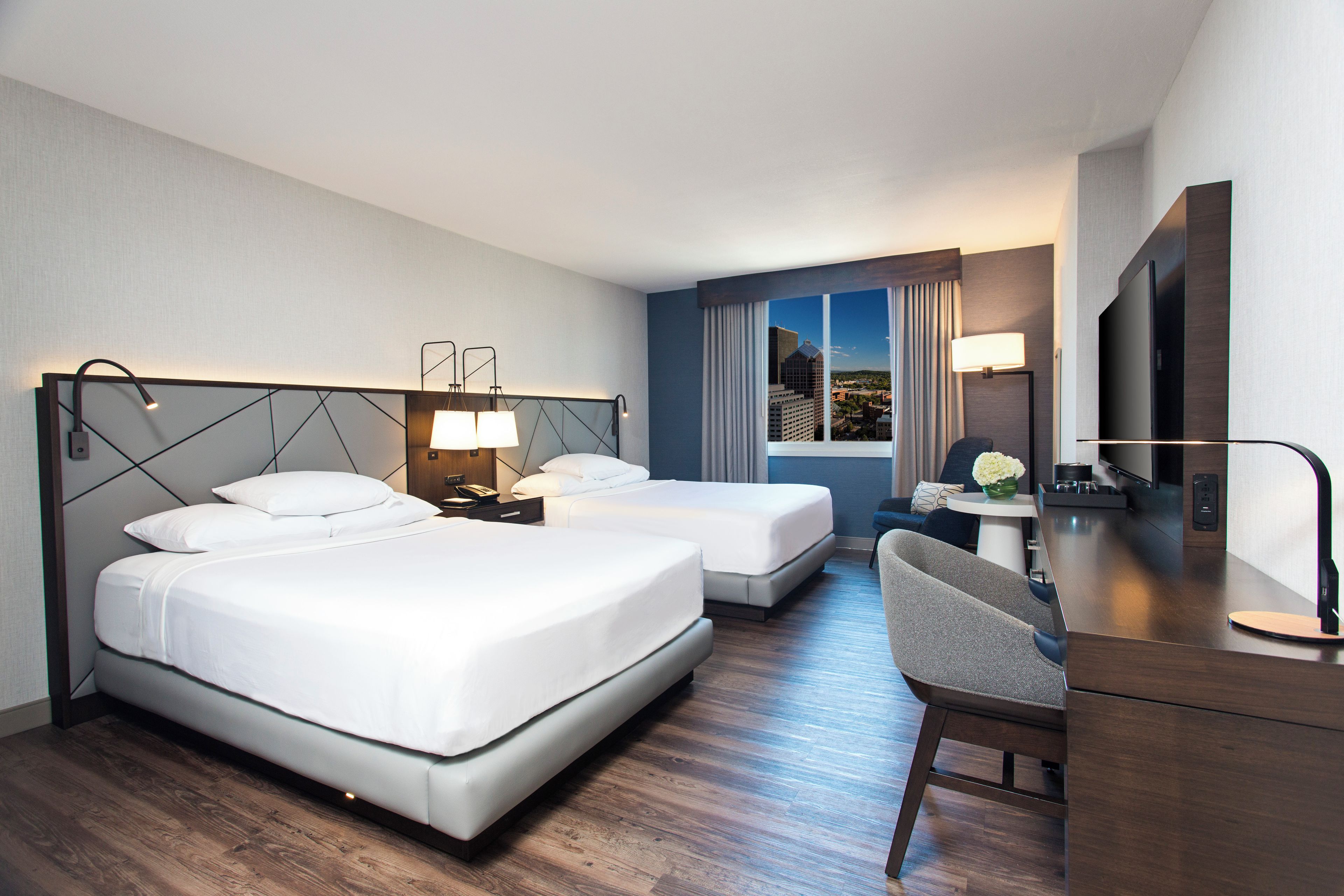 A hotel room with two beds, a desk, a chair, a TV, and a window with a city view.