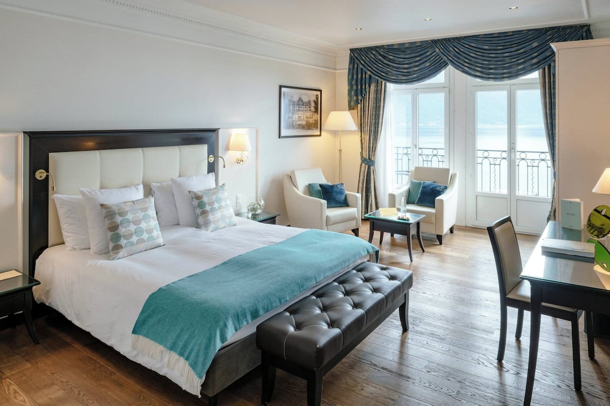 Una habitación de hotel con una cama grande, una zona de estar con una silla y una mesa, y un balcón con barandilla.