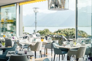 Un restaurante con mesas dispuestas para comer, amplios ventanales con vistas a un lago y montañas, y un diseño interior moderno.