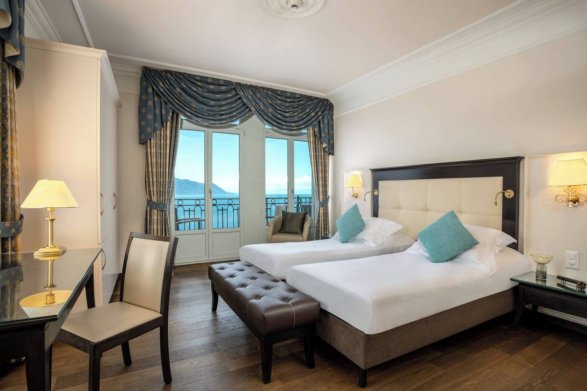 Habitación de hotel con dos camas, una silla, un banco, un escritorio con lámpara y vistas al mar.