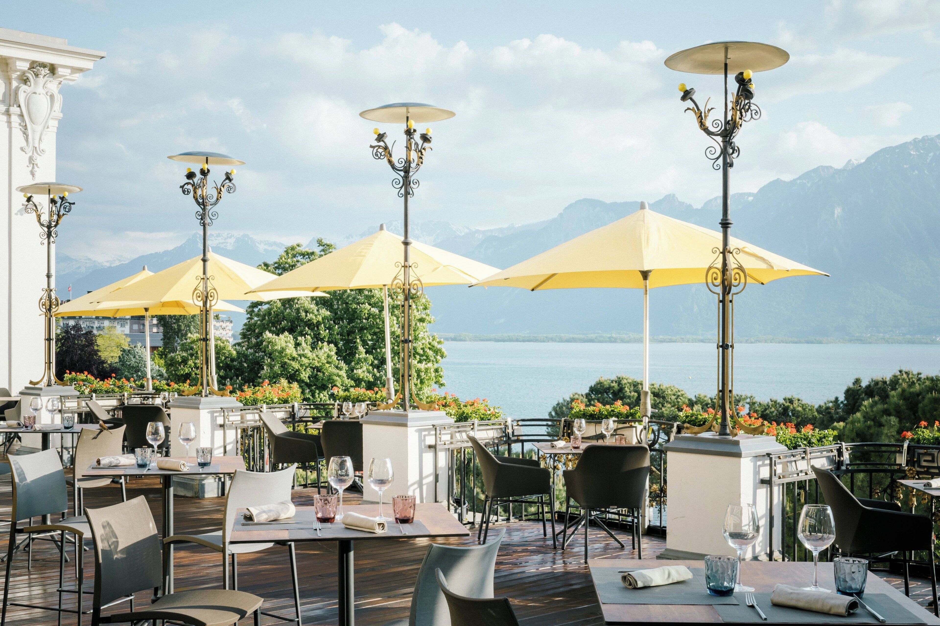 An outdoor dining area with tables, chairs, and yellow umbrellas overlooking a lake and mountains.
