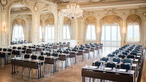 Un salón imponente con techo ricamente decorado y candelabros, filas de mesas y sillas, y amplias ventanas.