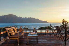 Una terraza de madera con mobiliario de mimbre, una mesa de vidrio y vistas a montañas y una masa de agua.