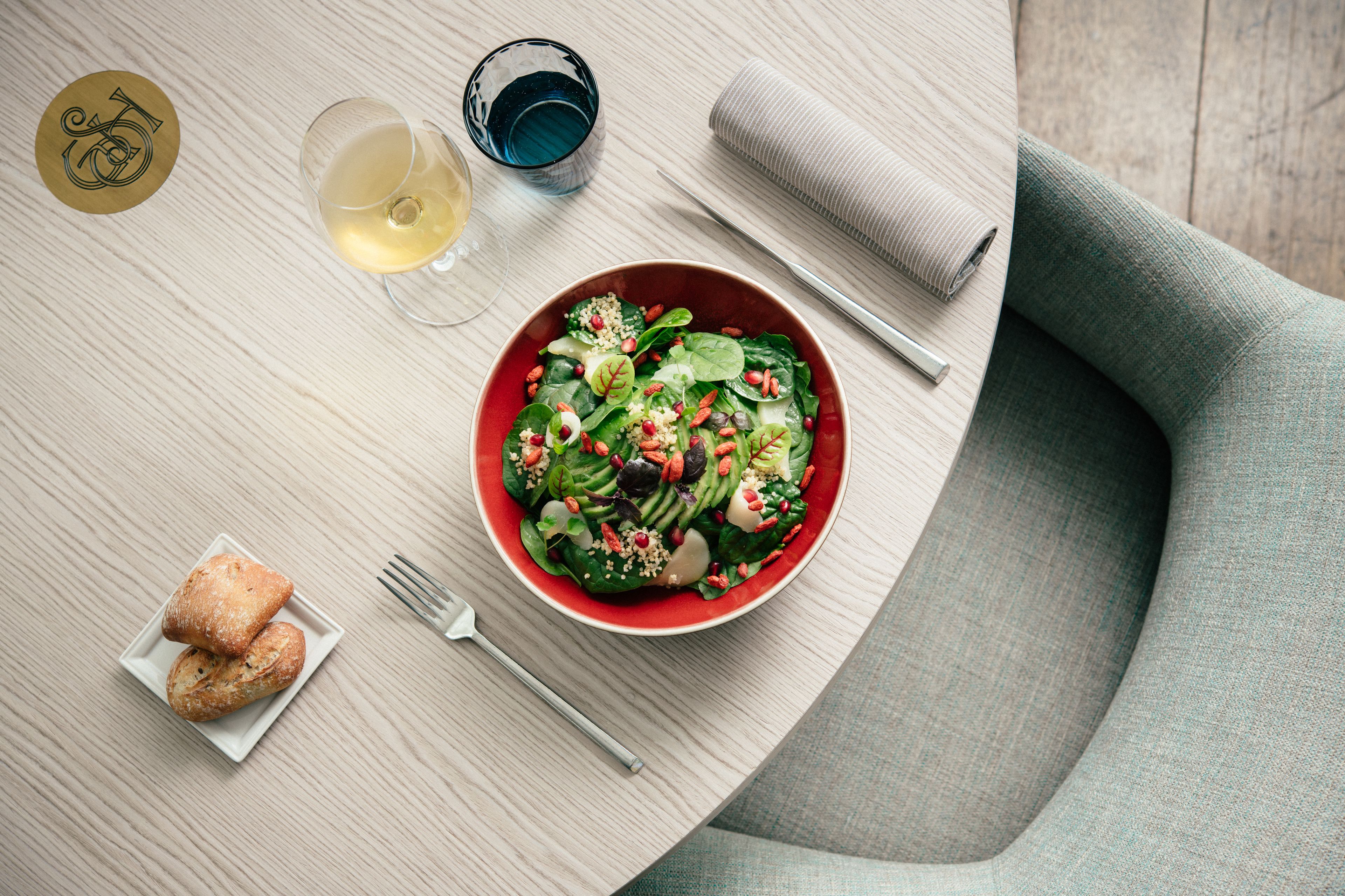 Une table avec un saladier rouge rempli de salade, deux verres de vin blanc, une petite assiette de pâtisseries et une serviette.