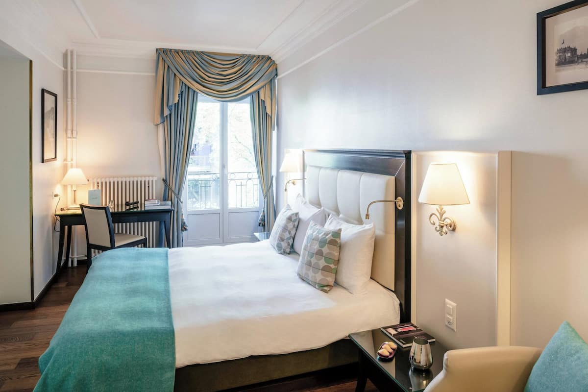 Habitación de hotel con una cama grande, un escritorio, una silla y vistas al exterior a través de una ventana con cortinas.