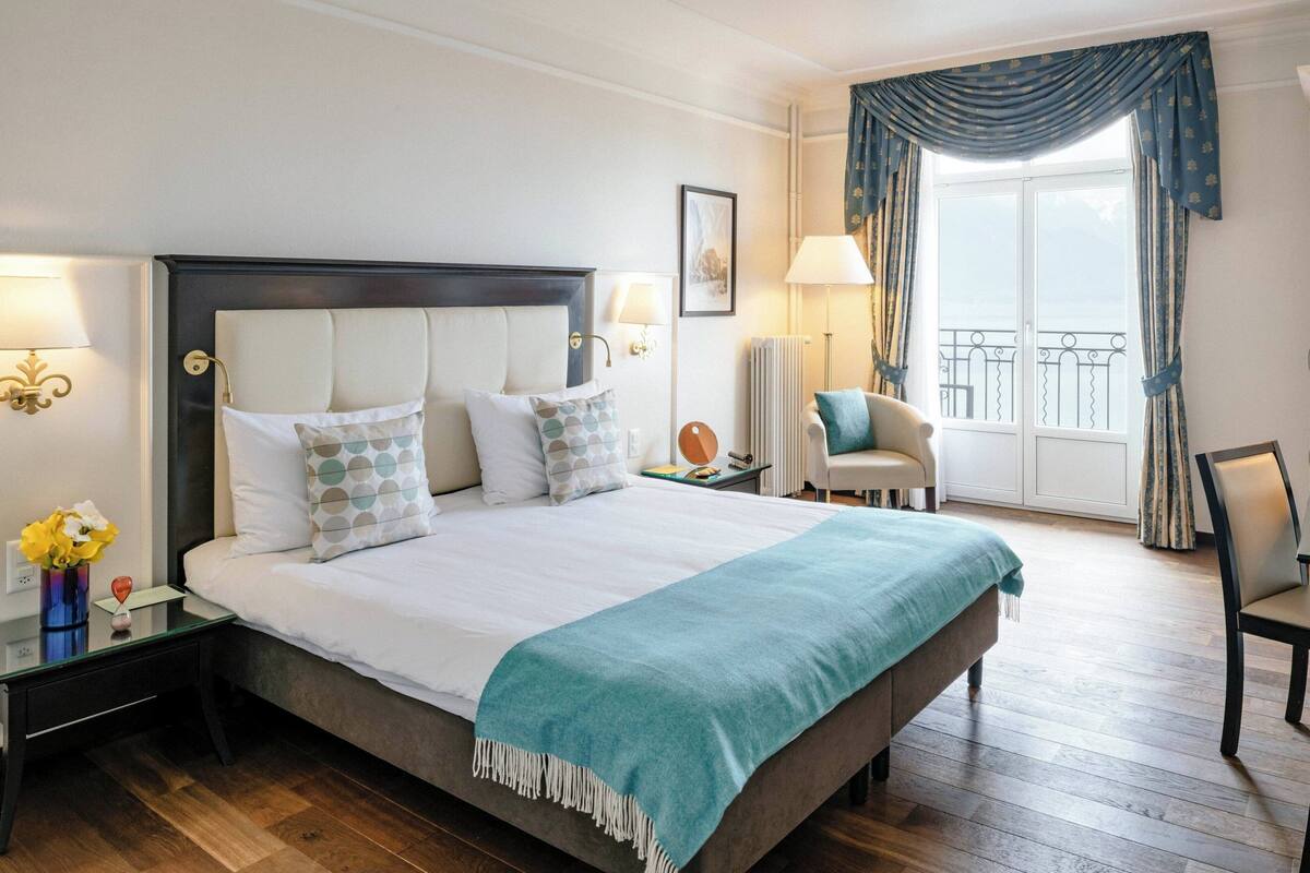 Habitación de hotel con una cama grande, dos mesitas de noche, una silla, una ventana con cortinas y vistas al exterior.