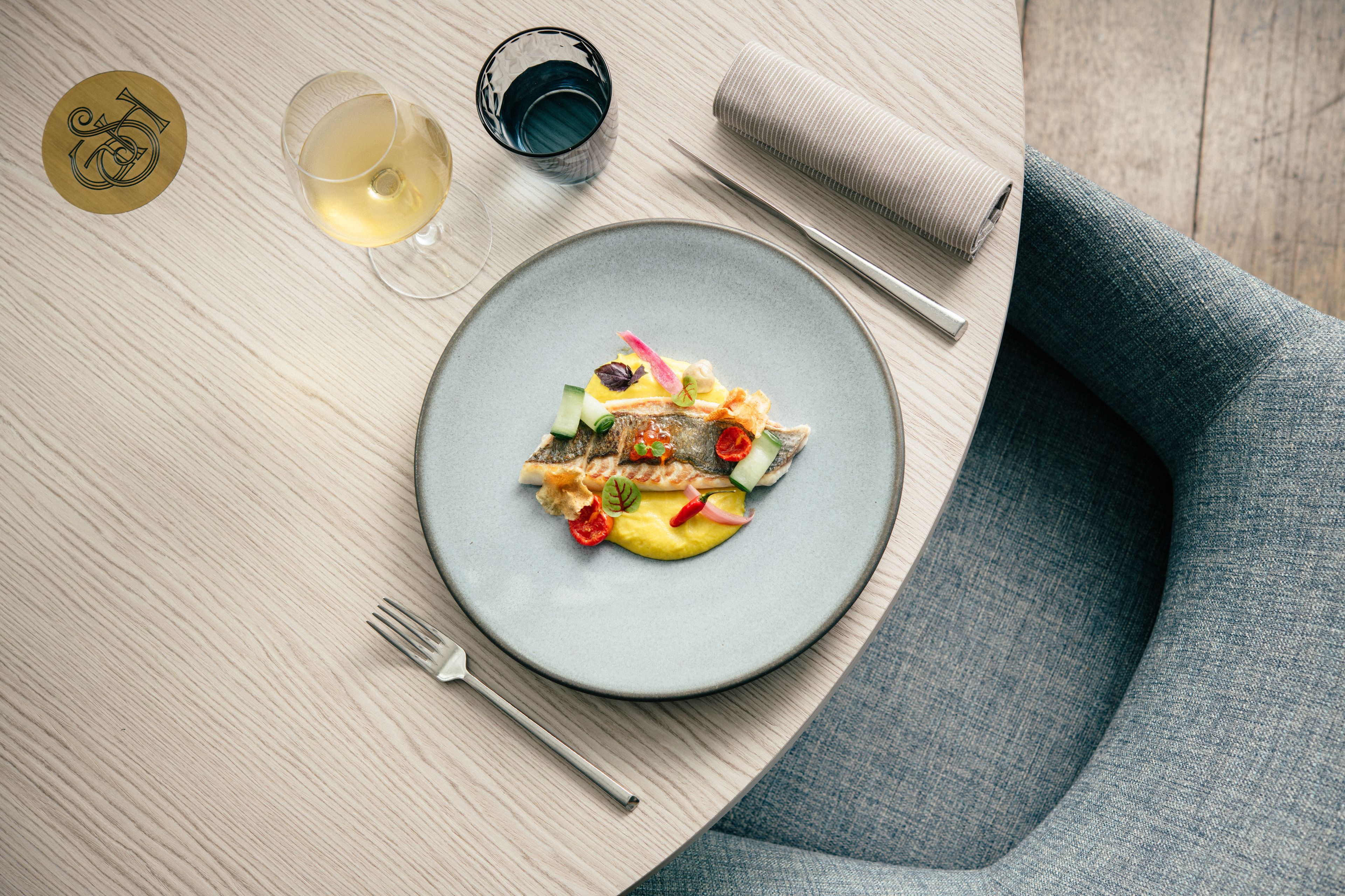 A dining table with a plate of food, a fork, a glass of white wine, and a napkin.
