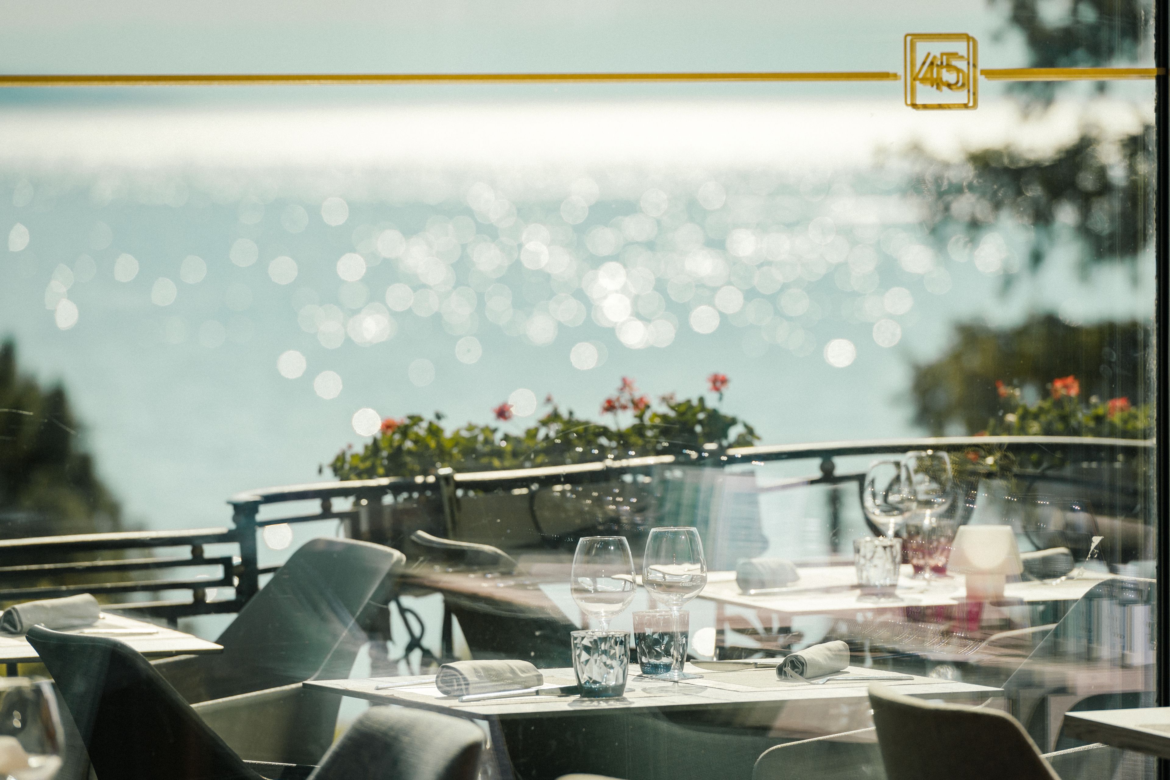 A dining area with tables set for two, a view of the sea, and a reflection of the sky and flowers on the window.