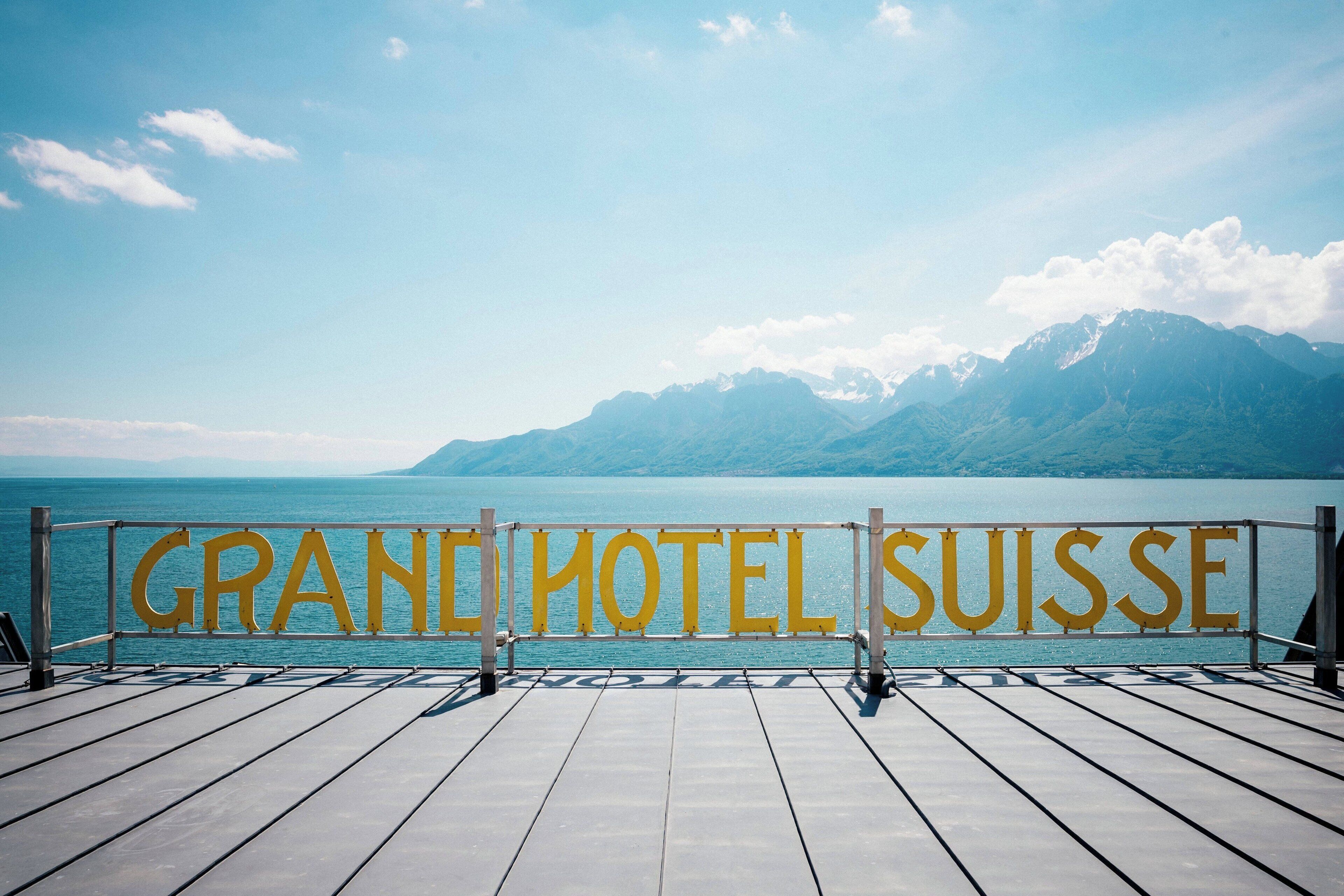 Eine Holzterrasse mit einem Schild, auf dem 'GRAND HOTEL SUISSE' steht, mit Blick auf ein Gewässer und Berge.
